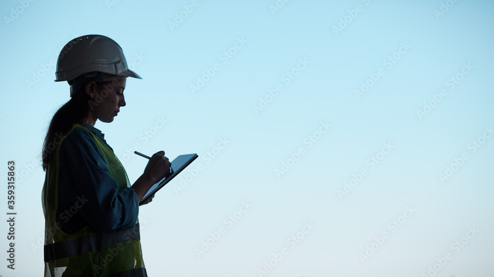 Wall mural silhouette of an engineer in a hard hat and holding a tablet against the blue sky