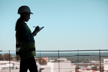 silhouette of an engineer in a hard hat and with a tablet in his hands against the blue sky and the city
