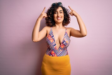 Young beautiful arab woman on vacation wearing swimsuit and sunglasses over pink background smiling pointing to head with both hands finger, great idea or thought, good memory