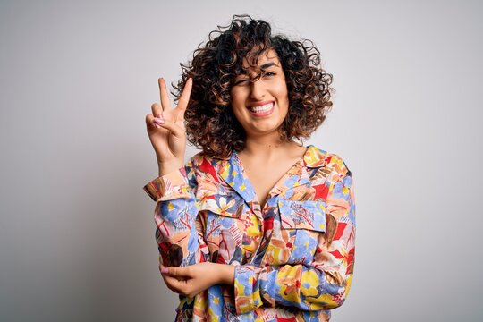 Young beautiful curly arab woman wearing floral colorful shirt standing over white background smiling with happy face winking at the camera doing victory sign. Number two.