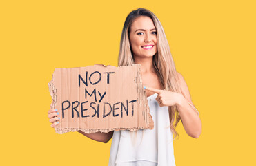 Young beautiful blonde woman holding not my president cardboard banner smiling happy pointing with hand and finger