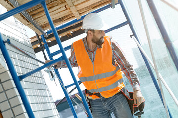 Fototapeta na wymiar A stylish Builder with a beard in an orange vest with a safety belt conducts construction work