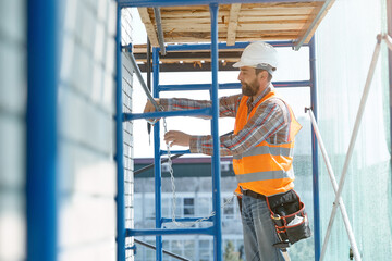 Fototapeta na wymiar A stylish Builder with a beard in an orange vest with a safety belt conducts construction work