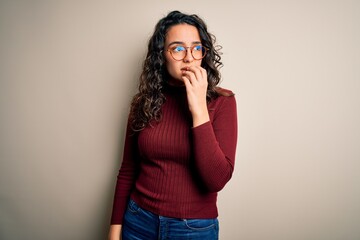 Beautiful woman with curly hair wearing casual sweater and glasses over white background looking stressed and nervous with hands on mouth biting nails. Anxiety problem.