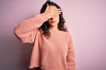 Young beautiful woman with curly hair wearing casual sweater over isolated pink background covering eyes with hand, looking serious and sad. Sightless, hiding and rejection concept