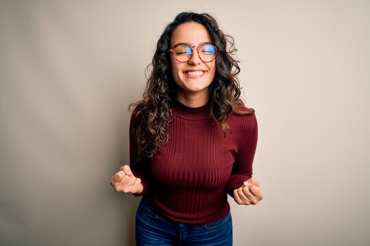 Beautiful woman with curly hair wearing casual sweater and glasses over white background very happy and excited doing winner gesture with arms raised, smiling and screaming for success. Celebration