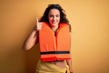 Young beautiful woman with curly hair wearing orange lifejacket over yellow background doing happy...