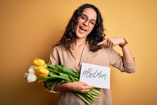 Young Beautiful Mom With Curly Hair Holding Flowers And Card As Mothers Day Present With Surprise Face Pointing Finger To Himself