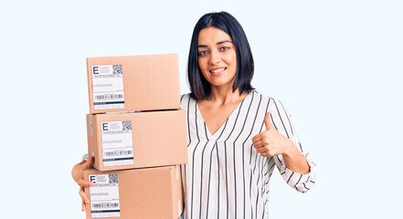 Young beautiful latin woman holding delivery package smiling happy and positive, thumb up doing excellent and approval sign
