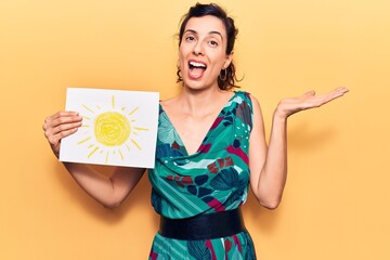 Young beautiful hispanic woman holding sun draw celebrating achievement with happy smile and winner expression with raised hand