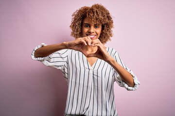 Beautiful african american woman with curly hair wearing striped t-shirt over pink background smiling in love doing heart symbol shape with hands. Romantic concept.
