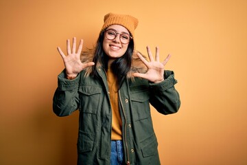 Young brunette woman wearing glasses and winter coat with hat over yellow isolated background showing and pointing up with fingers number ten while smiling confident and happy.