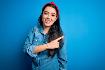 Young brunette woman wearing casual denim shirt over blue isolated background cheerful with a smile on face pointing with hand and finger up to the side with happy and natural expression