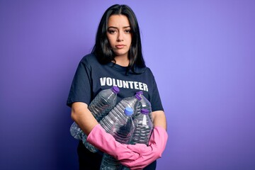 Young brunette woman wearing volunteer t-shirt picking plastic bottles over purple background with a confident expression on smart face thinking serious