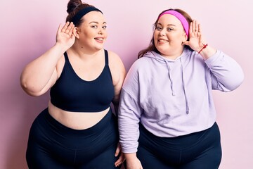 Young plus size twins wearing sportswear smiling with hand over ear listening an hearing to rumor or gossip. deafness concept.