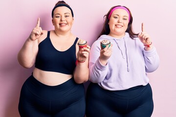 Young plus size twins wearing sportswear holding cupcake smiling with an idea or question pointing finger with happy face, number one