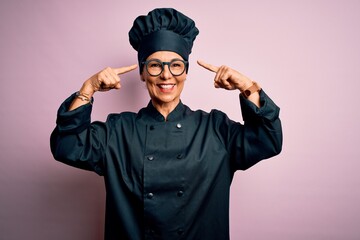 Middle age brunette chef woman wearing cooker uniform and hat over isolated pink background smiling pointing to head with both hands finger, great idea or thought, good memory