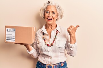 Senior grey-haired woman holding delivery package pointing thumb up to the side smiling happy with...