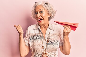 Senior grey-haired woman holding paper airplane pointing thumb up to the side smiling happy with open mouth