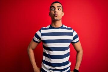 Handsome african american man wearing casual striped t-shirt standing over red background making fish face with lips, crazy and comical gesture. Funny expression.