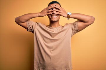 Young handsome african american man wearing casual t-shirt standing over yellow background covering eyes with hands smiling cheerful and funny. Blind concept.