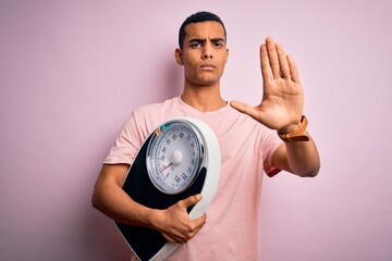 Young handsome african american slim sportsman using weight machine over pink background with open hand doing stop sign with serious and confident expression, defense gesture