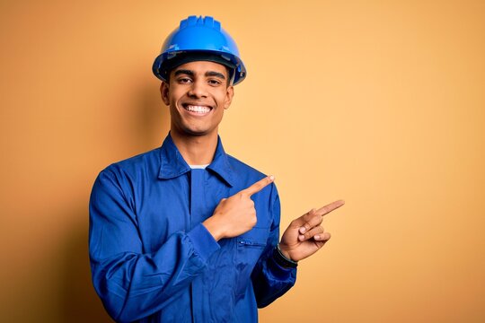Young handsome african american worker man wearing blue uniform and security helmet smiling and looking at the camera pointing with two hands and fingers to the side.