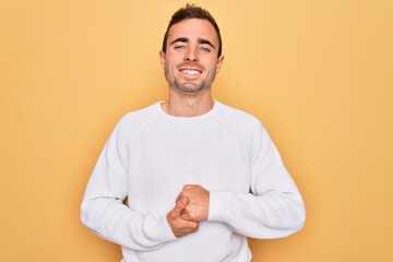 Young handsome man with blue eyes wearing casual sweater standing over yellow background with hands together and crossed fingers smiling relaxed and cheerful. Success and optimistic