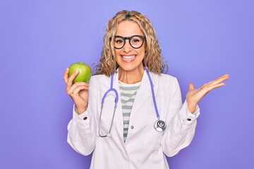 Young beautiful blonde doctor woman wearing stethoscope holding green apple fruit celebrating victory with happy smile and winner expression with raised hands