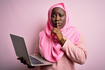 African american plus size woman wearing muslim hijab using laptop over pink background asking to be quiet with finger on lips. Silence and secret concept.
