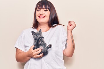 Young plus size woman holding cat screaming proud, celebrating victory and success very excited with raised arm
