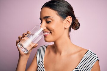 Young beautiful brunette woman smiling happy and confident. Standing with smile on face drinking glass of healthy water to refreshment over isolated pink background