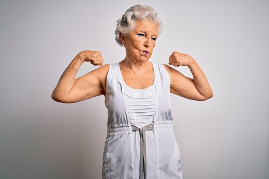 Senior beautiful grey-haired woman wearing casual summer dress over white background showing arms muscles smiling proud. Fitness concept.