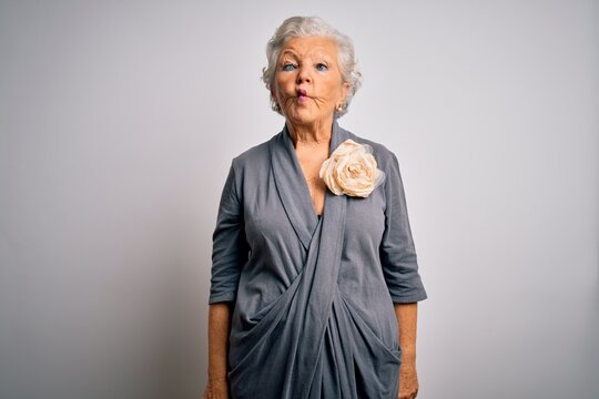 Senior beautiful grey-haired woman wearing casual dress standing over white background making fish face with lips, crazy and comical gesture. Funny expression.
