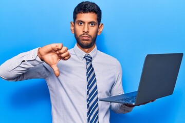 Young latin man holding laptop with angry face, negative sign showing dislike with thumbs down,...