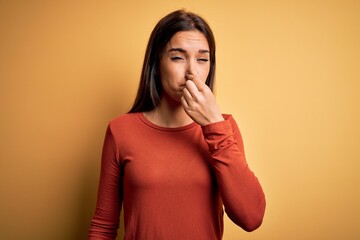 Young beautiful brunette woman wearing casual t-shirt standing over yellow background smelling something stinky and disgusting, intolerable smell, holding breath with fingers on nose. Bad smell - obrazy, fototapety, plakaty