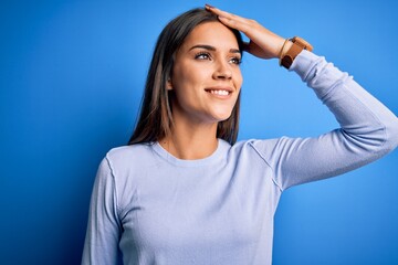 Fototapeta premium Young beautiful brunette woman wearing casual sweater standing over blue background smiling confident touching hair with hand up gesture, posing attractive and fashionable