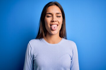 Young beautiful brunette woman wearing casual sweater standing over blue background sticking tongue...