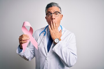 Middle age handsome grey-haired doctor man holding pink cancer ribbon cover mouth with hand shocked with shame for mistake, expression of fear, scared in silence, secret concept