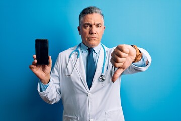 Middle age handsome grey-haired doctor man holding smartphone showing screen with angry face, negative sign showing dislike with thumbs down, rejection concept
