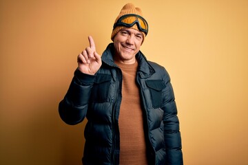 Middle age handsome grey-haired skier man on vacation wearing ski goggles showing and pointing up with finger number one while smiling confident and happy.