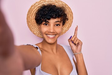 Beautiful african american tourist woman wearing bikini and hat making selfie by camera surprised with an idea or question pointing finger with happy face, number one