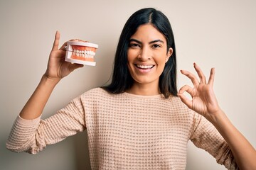 Young beautiful hispanic dentist woman holding prosthesis dental mouth doing ok sign with fingers, excellent symbol