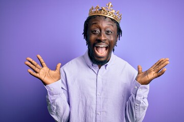 Young african american man wearing golden crown of king over isolated purple background celebrating crazy and amazed for success with arms raised and open eyes screaming excited. Winner concept