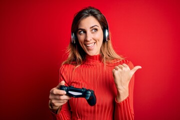 Young beautiful brunette gamer woman playing video game using joystic and headphones pointing and showing with thumb up to the side with happy face smiling