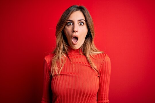 Young beautiful brunette woman wearing casual turtleneck sweater over red background afraid and shocked with surprise and amazed expression, fear and excited face.