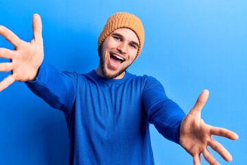 Naklejka premium Young handsome man wearing casual sweater and wool cap looking at the camera smiling with open arms for hug. cheerful expression embracing happiness.