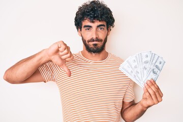 Handsome young man with curly hair and bear holding money bag with dollar symbol with angry face,...