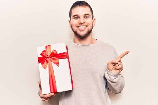 Young handsome man holding gift smiling happy pointing with hand and finger to the side