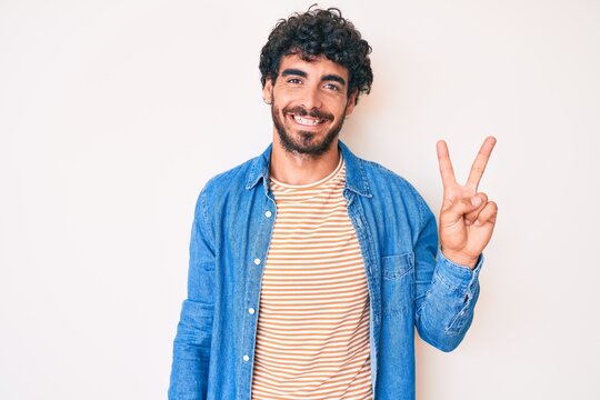 Handsome young man with curly hair and bear wearing casual denim jacket showing and pointing up with fingers number two while smiling confident and happy.